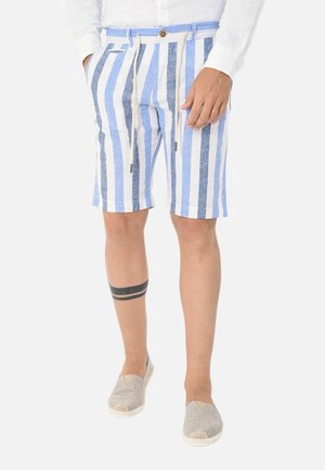 Uomo che indossa pantaloncini a righe verticali blu e bianche, una camicia bianca a maniche lunghe, scarpe slip-on grigie e un tatuaggio a doppia fascia nera sul polpaccio sinistro.