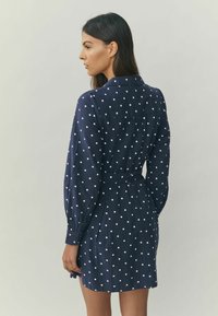 Marineblaues Kleid mit weißen Polka Dots, langen Ärmeln, Kragen, tailliert und einem plissierten Rock. Strukturierter Stoff, midilang bis zur Mitte des Oberschenkels.