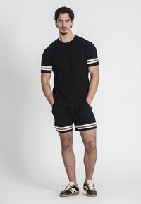 T-shirt in maglia nera a costine con accenti a strisce bianche e marroni sulle maniche, abbinato a pantaloni corti coordinati con strisce simili. Le sneakers completano il look.