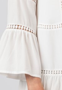 Vit blus med utställda ärmar och lager av volanger. Den har intrikata spetsdetaljer längs sömmarna och en mjuk, flödande textur.