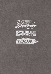 Šedá tkanina s bílým tištěným textem: "USA EXTREME MOTO," "MOTO RACING" a "RACING TOUR," s texturedním, opotřebovaným vzhledem.