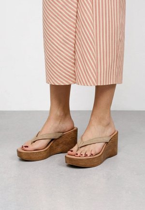 Pies con chanclas beige de cuña con suelas de plataforma de madera y pantalones cortos a rayas rosas y blancos.