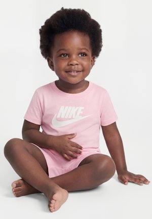 Bambino piccolo con capelli ricci seduto a gambe incrociate, sorridente, indossa una maglietta rosa Nike e pantaloncini su sfondo bianco.