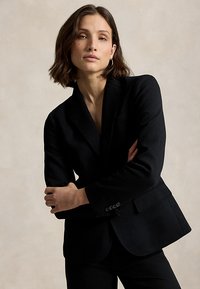 Kvinde med kort brunt hår iført en sort skræddersyet blazer og øreringe i ringform, stående foran en ensfarvet beige baggrund.
