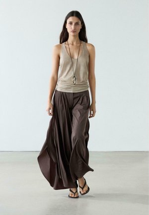 Frau geht vorwärts, trägt ein beiges ärmelloses Oberteil, fließende dunkelbraune weite Hosen, eine Anhängerkette und schwarze Sandalen auf einem hellen Boden.