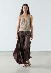 Femme marchant vers l'avant portant un haut sans manches beige, un pantalon ample à jambes larges marron foncé fluide, un collier pendentif et des sandales noires sur un sol clair.