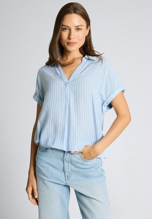 Femme aux longs cheveux bruns portant un chemisier rayé bleu clair à manches courtes et un jean bleu clair, debout avec une main dans la poche.