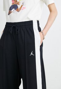 Pantalons de sport noirs avec une bande latérale blanche, une taille élastique avec cordon de serrage et un logo Jordan blanc sur la jambe gauche.