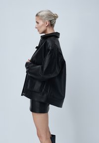 Veste en cuir noir à coupe décontractée, avec un grand col et des détails de couture visibles. Texture lisse et poches latérales.