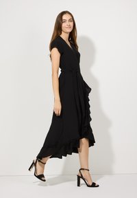 Schwarzes Kleid mit kurzen Ärmeln, Wickeldesign, Rüschensaum und Gürteldetail. Kombiniert mit schwarzen Absatzsandalen mit goldenen Akzenten.