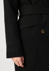 Abrigo de lana negro con un cinturón ancho, cierre de botones y un bolsillo lateral. La textura parece suave con un diseño estructurado.