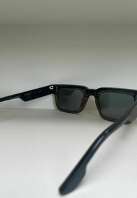 Zwarte en bruine rechthoekige zonnebril met donkere lenzen. Kenmerkt een matte afwerking van het frame en een onderscheidend geometrisch ontwerp. Fabrikantgegevens gelabeld.