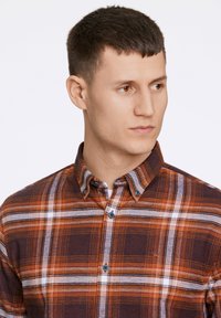 Camicia a quadri marrone con righe arancioni e bianche, colletto button-down e bottoni blu. Il materiale sembra avere una texture con una vestibilità aderente.