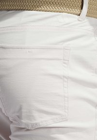 Los pantalones de algodón blancos presentan un bolsillo trasero con costuras sutiles y una textura suave. Un cinturón tejido beige complementa la cintura.