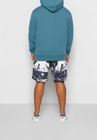 Hombre con sudadera color teal, pantalones cortos de estampado abstracto en blanco y negro, y zapatillas blancas con detalles en azul y rojo, de pie contra un fondo liso.