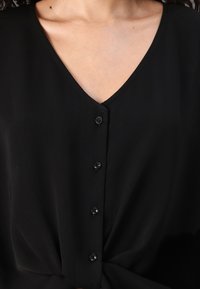 Juoda bliuselė su V formos iškirpte, turinti šešis mažus mygtukus priekyje, surišimo detalę liemens srityje ir švelnų, krentančio audinio tekstūrą.