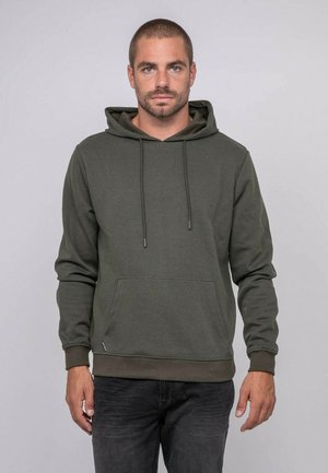 Homme aux cheveux courts et à la barbe portant un sweat à capuche vert olive et un jean foncé, debout devant un fond clair uni.