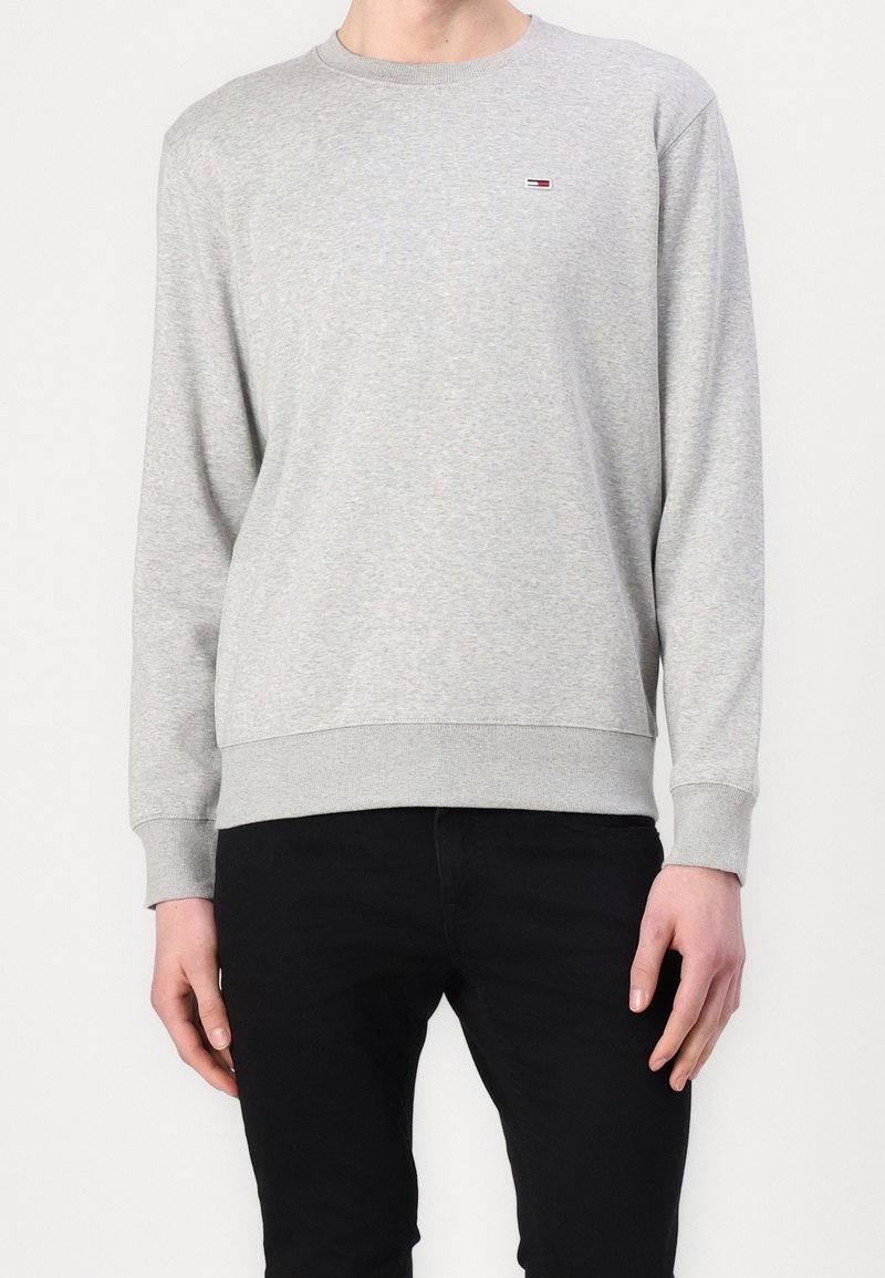Homme portant un sweat-shirt gris clair à manches longues et col rond avec un petit logo drapeau sur la poitrine et un pantalon noir, debout les bras détendus.