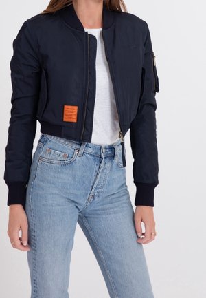 Femme portant une veste bomber noire courte avec un patch marron et un jean taille haute bleu clair, sur un fond blanc uni.
