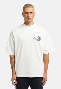 Witte katoenen T-shirt met korte raglanmouwen, voorzien van een kleine geprinte afbeelding van een vis en cirkelvormige tekst op de linkerkant van de borst.