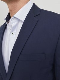 Blazer marine à coupe ajustée, en tissu lisse. Le col présente une chemise habillée à motif bleu clair, visible en dessous.