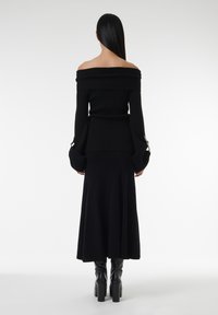 Haut en maille noire à épaules dénudées avec des manches larges, associé à une jupe noire évasée. La tenue présente une taille cintrée et une texture lisse.