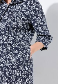 Abito a stampa floreale in blu navy con rose bianche; presenta un colletto, maniche arrotolate e tasche; realizzato in un tessuto morbido con una texture liscia.