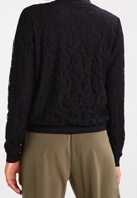 Giacca in pizzo nero con motivo floreale testurizzato, maniche lunghe e cintura aderente in vita. La parte posteriore mostra dettagli elaborati in pizzo.