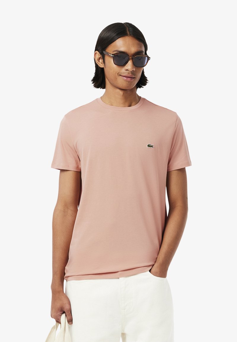 Světle růžové tričko s kulatým výstřihem vyrobené z měkké tkaniny, s malým zeleným logem Lacoste na hrudi. Model nosí tmavé sluneční brýle.