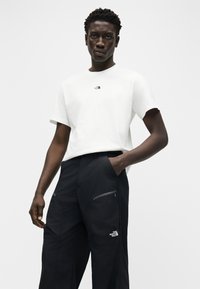 Άνδρας που φοράει λευκό T-shirt The North Face και μαύρο παντελόνι με εμπρόσθιες τσέπες με φερμουάρ, στέκεται με το ένα χέρι στην τσέπη, μπροστά σε απλό φόντο.