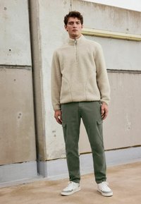 Pull en polaire beige avec un zip demi-long, doté de poches latérales. Associé à un pantalon cargo vert olive et des baskets blanches. Texture douce.