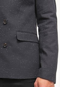 Czarna marynarka w białe kropki z wycięciem na klapę, dwoma guzikami i pojedynczą kieszenią na piersi. Tkanina ma fakturowane wykończenie z widocznymi białymi plamkami.