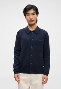 Σκούρο μπλε πλεκτό πουλόβερ από υφασμάτινη υφή, με γιακά, κλείσιμο με κουμπί και τσέπη στο στήθος. Μακριά μανίκια με λεπτομέρειες κουμπιών.