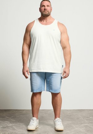 Man standing, wearing a white sleeveless tank top, light blue denim shorts, and white sneakers on a gray floor.