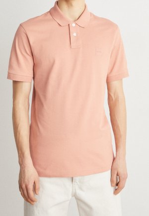 Homme portant un polo à manches courtes rose clair avec deux boutons blancs et un pantalon blanc sur un fond uni.
