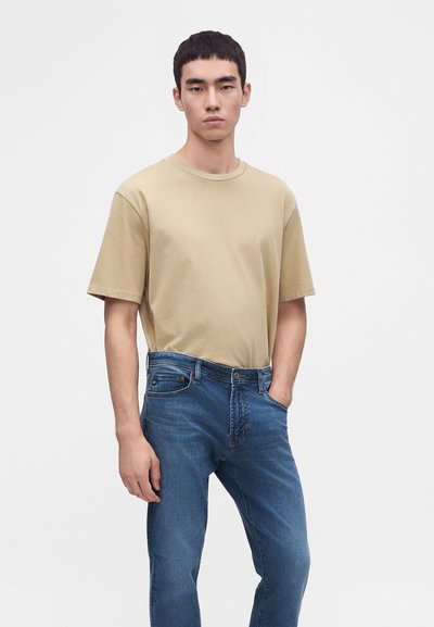 Jeune homme debout portant un t-shirt beige à manches courtes et un jean bleu, une main dans la poche, sur un fond clair uni.