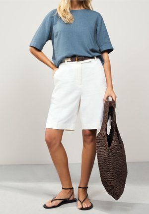 Woman wearing blue short-sleeve top, white knee-length shorts with brown belt, black sandals, and holding large brown woven bag.