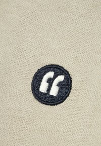 Bežinė medžiaga su apskritu tamsiai mėlynos spalvos siuvinėtu ženkleliu centre, aplink kurį yra baltos kabutės. Lygi tekstūra, minimalus dizainas.