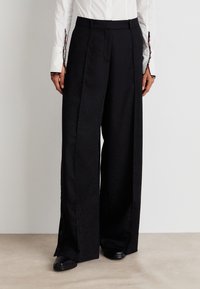 Pantalons noirs à jambe large avec une texture subtile, dotés de plis avant et de détails de boutons sur le côté. Portés avec une chemise blanche pour le contraste.