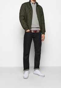 Kurtka pikowana w oliwkowym kolorze, szary sweter i koszula w kratę nałożone na siebie, w połączeniu z ciemnoniebieskimi dżinsami i białymi trampkami.