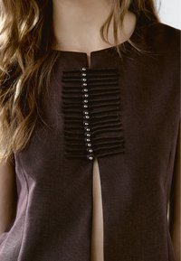 Bruine kleding met een ronde halslijn, verticale ruches met knopen in het midden en een subtiele split aan de onderkant. Geweven stof.