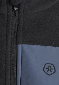 Giacca in pile con corpo nero e pannello anteriore grigio, dotata di zip frontale e patch con logo sul lato destro. Tessuto liscio.