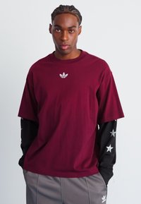 Bordeaux katoenen t-shirt met een ronde hals en korte mouwen, voorzien van een klein wit Adidas-logo op de borst en gelaagd met een zwart long-sleeve shirt.