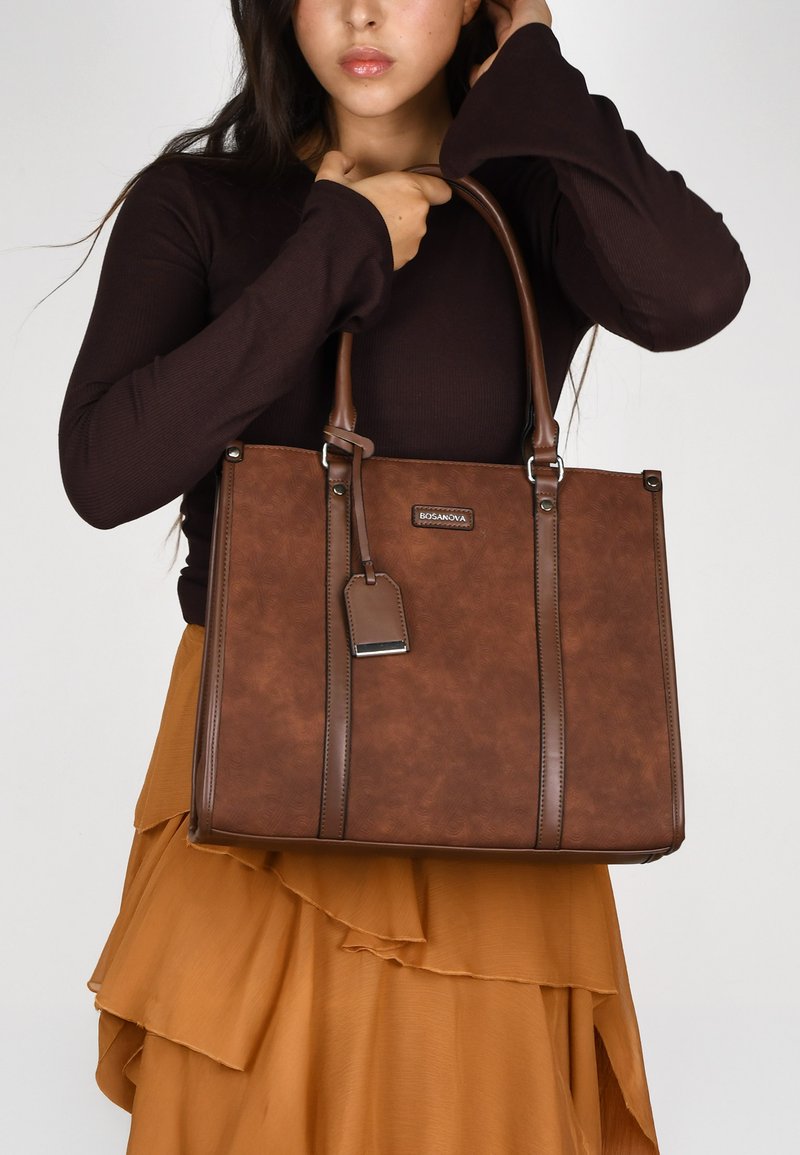 Bolso tote sintético marrón con asas dobles, superficie texturizada, detalle de logo en la parte delantera y una etiqueta desmontable, combinado con una falda en capas.