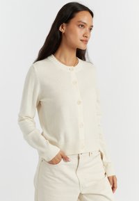 Cardigan crema realizzato in morbido materiale a maglia con scollatura tonda, chiusura con bottoni e polsini e orlo a coste.