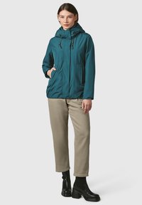 Giacca leggera color teal con cappuccio, chiusura con zip, cordino e tasche frontali. Indossata con chinos beige e stivali neri con tacco. Tessuto liscio.