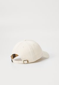 Beige bomulds-baseballkasket med en buet skygge, justerbar rem og et logomærke på bagsiden. Glat tekstur med syningsdetaljer.