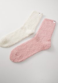 Par de meias macias e felpudas; uma em creme e a outra em rosa claro. Ambas têm uma textura fofinha e um design em cor sólida com uma pequena etiqueta de logotipo.