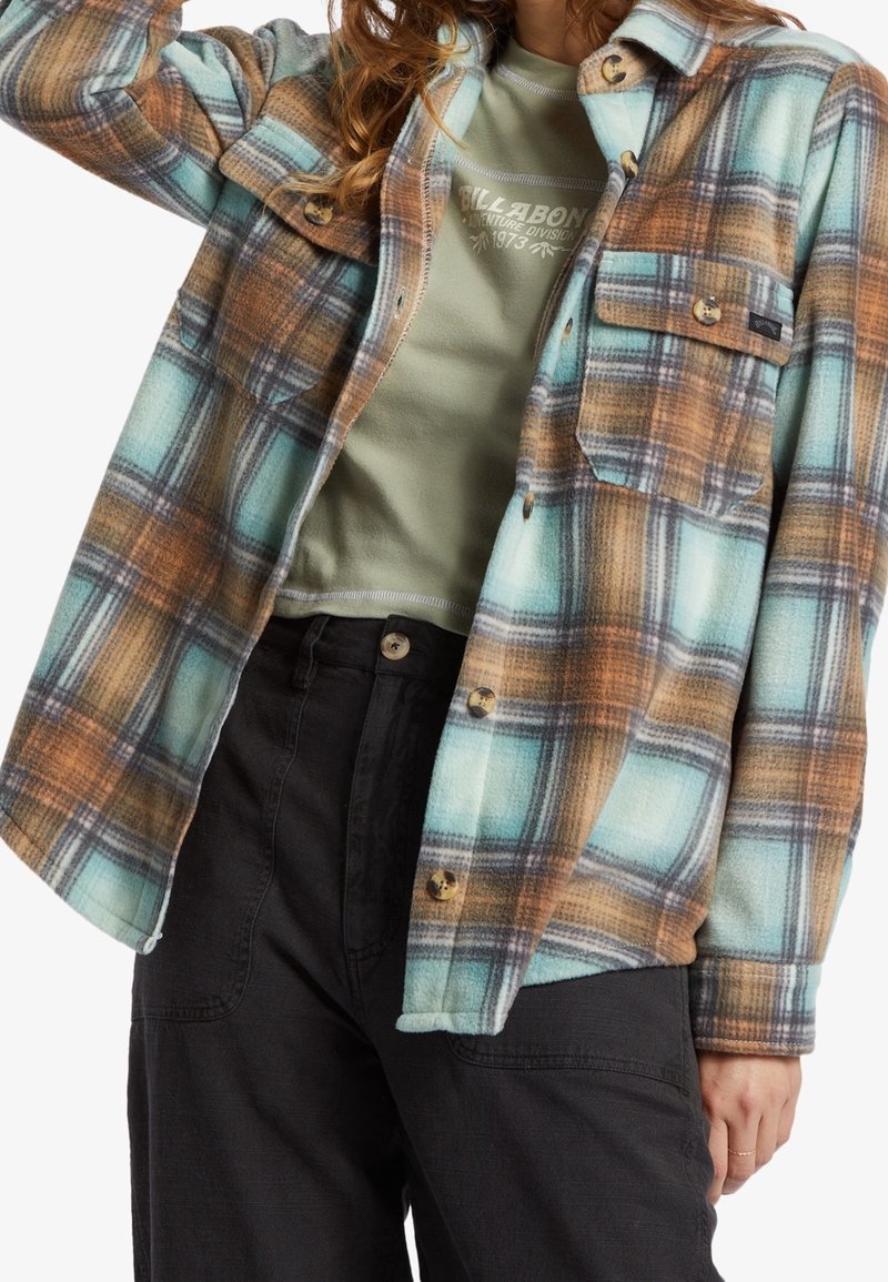 Personne portant une veste à carreaux bleue, marron et blanche boutonnée, par-dessus un t-shirt vert Billabong et un pantalon noir avec des boutons.