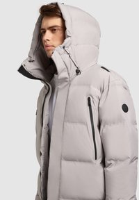 Duvet jakna u svijetlosivoj boji, s velikom kapuljačom, bočnim dzepovima na zip, i crnim detaljima. Glatka tkanina s kvadratnom teksturom.